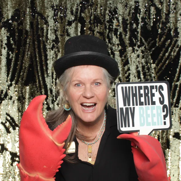 Close up of a guest in a bowler hat and lobster claw gloves holding a WHERE'S MY BEER sign